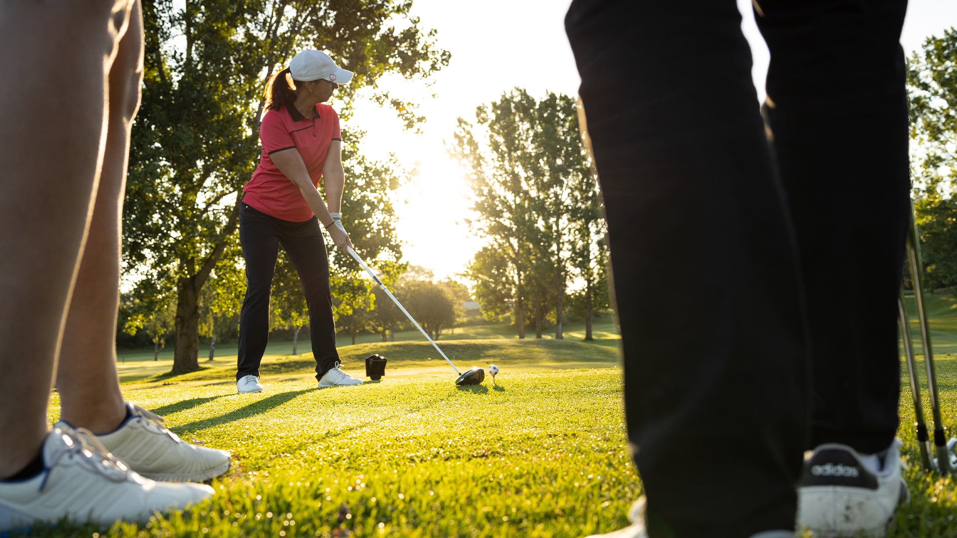 Kvinna i rosa tröja slå ut på tee. Golfvänner står bredvid, ser benen.